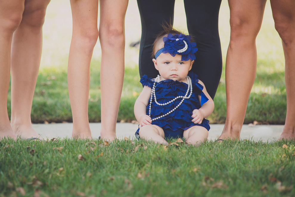 photography of baby wearing blue dress, headband and pearls sits unamused among legs of bridesmaids before a wedding in Buffalo, NY