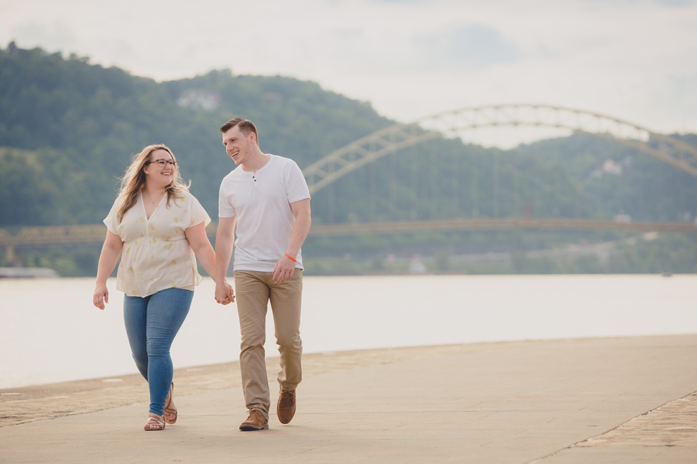 engagement photography of couple laughing and holding hands while walking in Point Park in Pittsburgh with bridge and river behind them