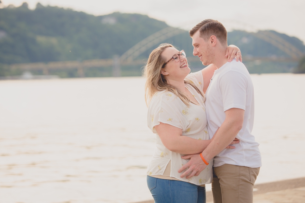 engagement photography of couple laughing in Point Park in Pittsburgh with bridge and river behind them