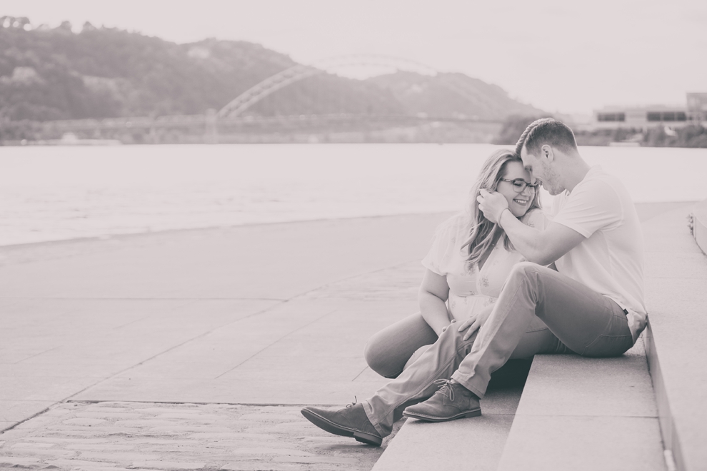 black and white engagement photography of couple sitting on steps in Point Park in Pittsburgh with bridge and river behind them