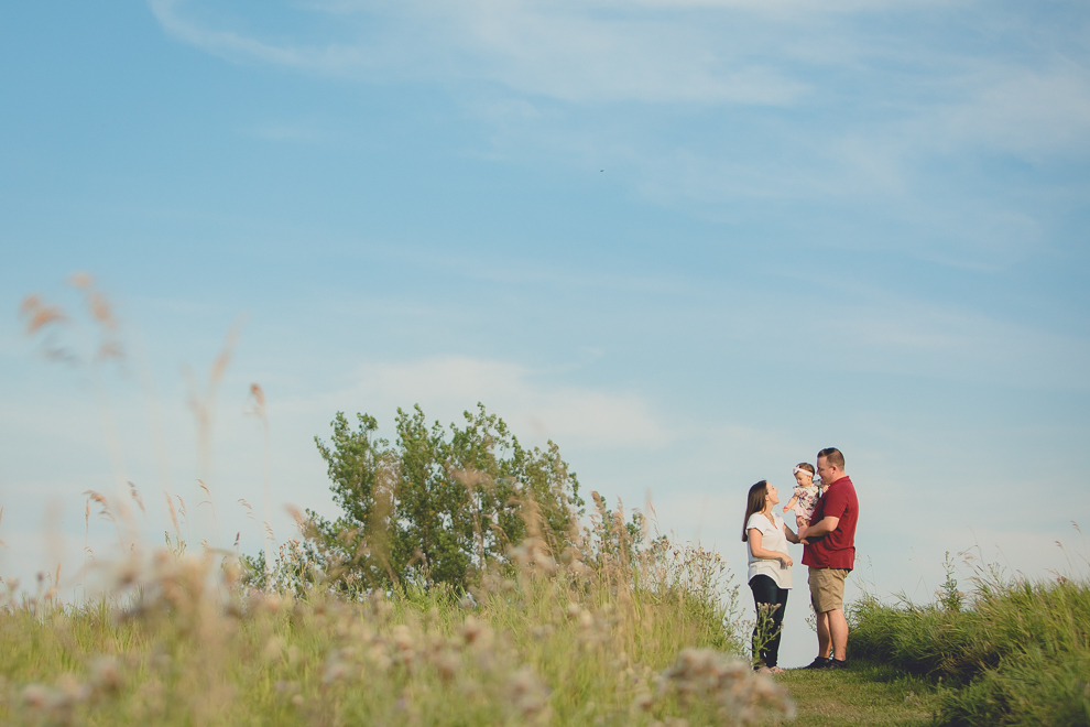 mom and dad stand with baby on top of hill in field during family portrait photography session in at Tifft Nature Preserve in Buffalo, NY