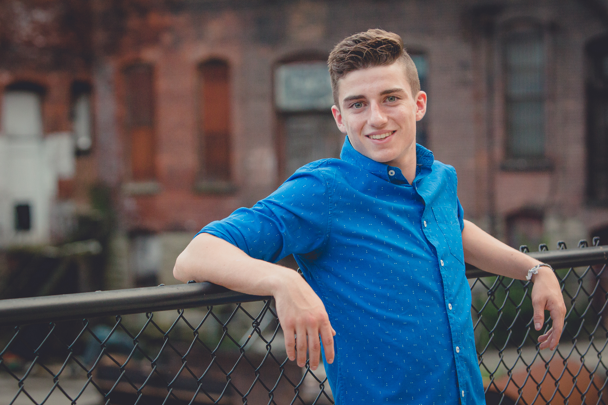 senior portrait of young man leaning on fence in front of brick wall in Jamestown during photography session 