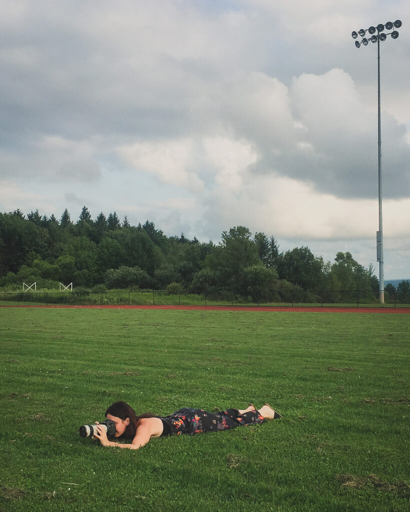 senior portrait photographer lays on stomach in grass on football field near Buffalo, NY to get perfect angle for shot 