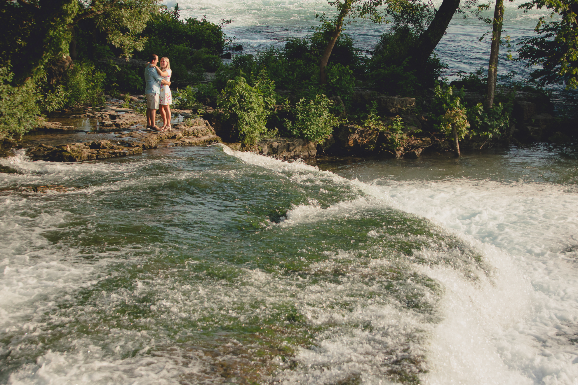 future bride and groom embrace in front of rapids on Goat Island during wedding engagement photography session in Niagara Falls State Park in NY