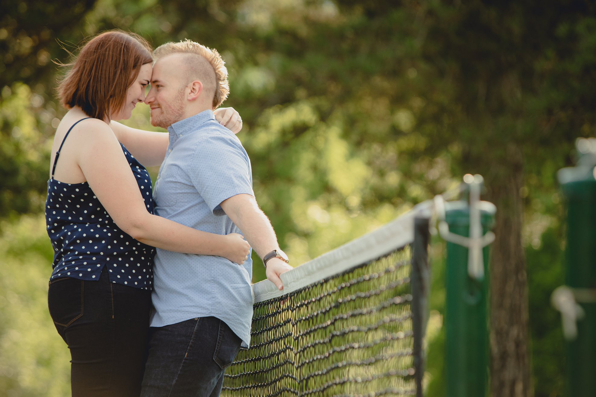 engaged couple poses for photographer with foreheads touching while leaning against a tennis net at Akron Falls Park near Buffalo, NY