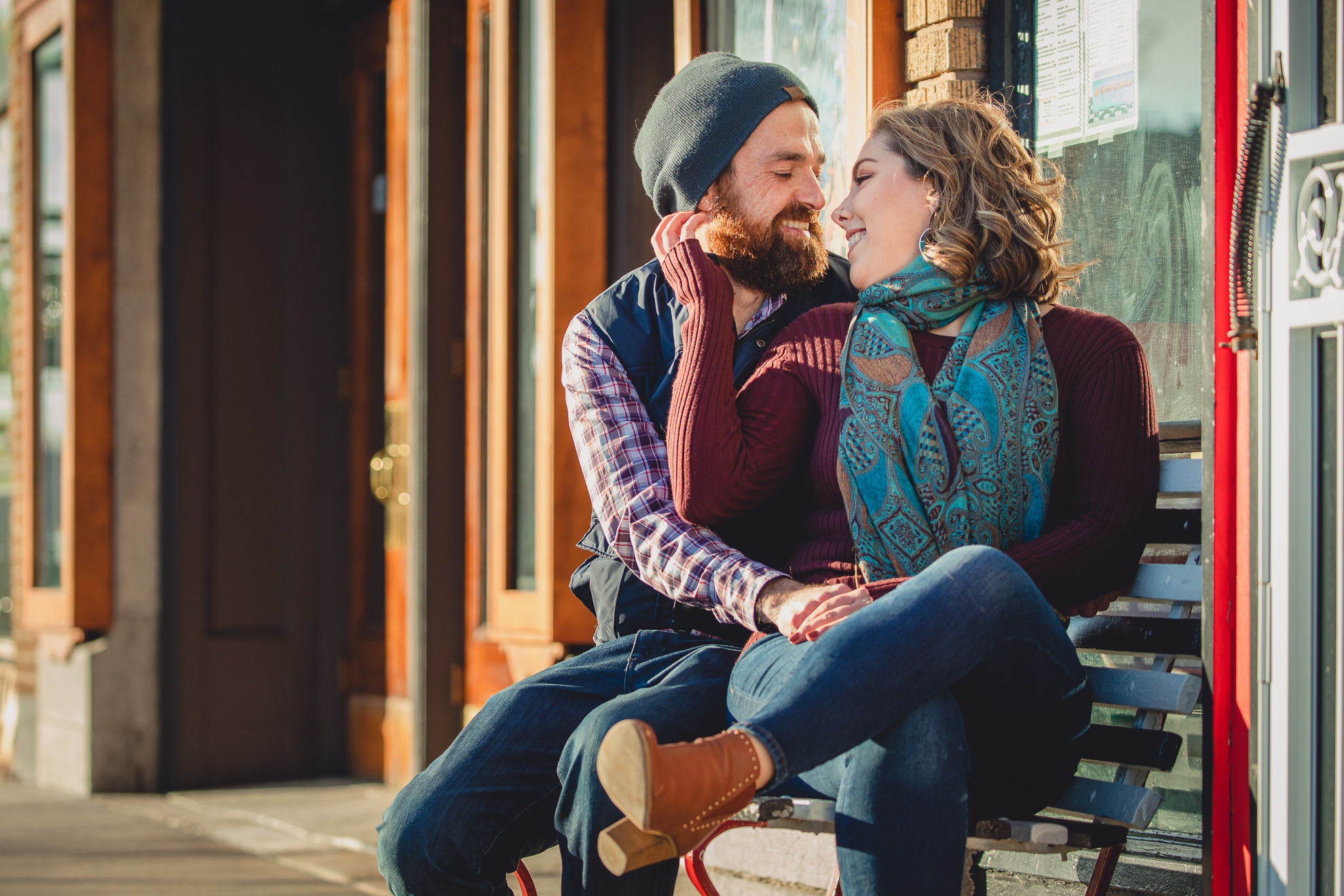 couple smiles at each other on bench in front of North Park Theater during couples mini photography session on Hertel Ave. in North Buffalo, NY