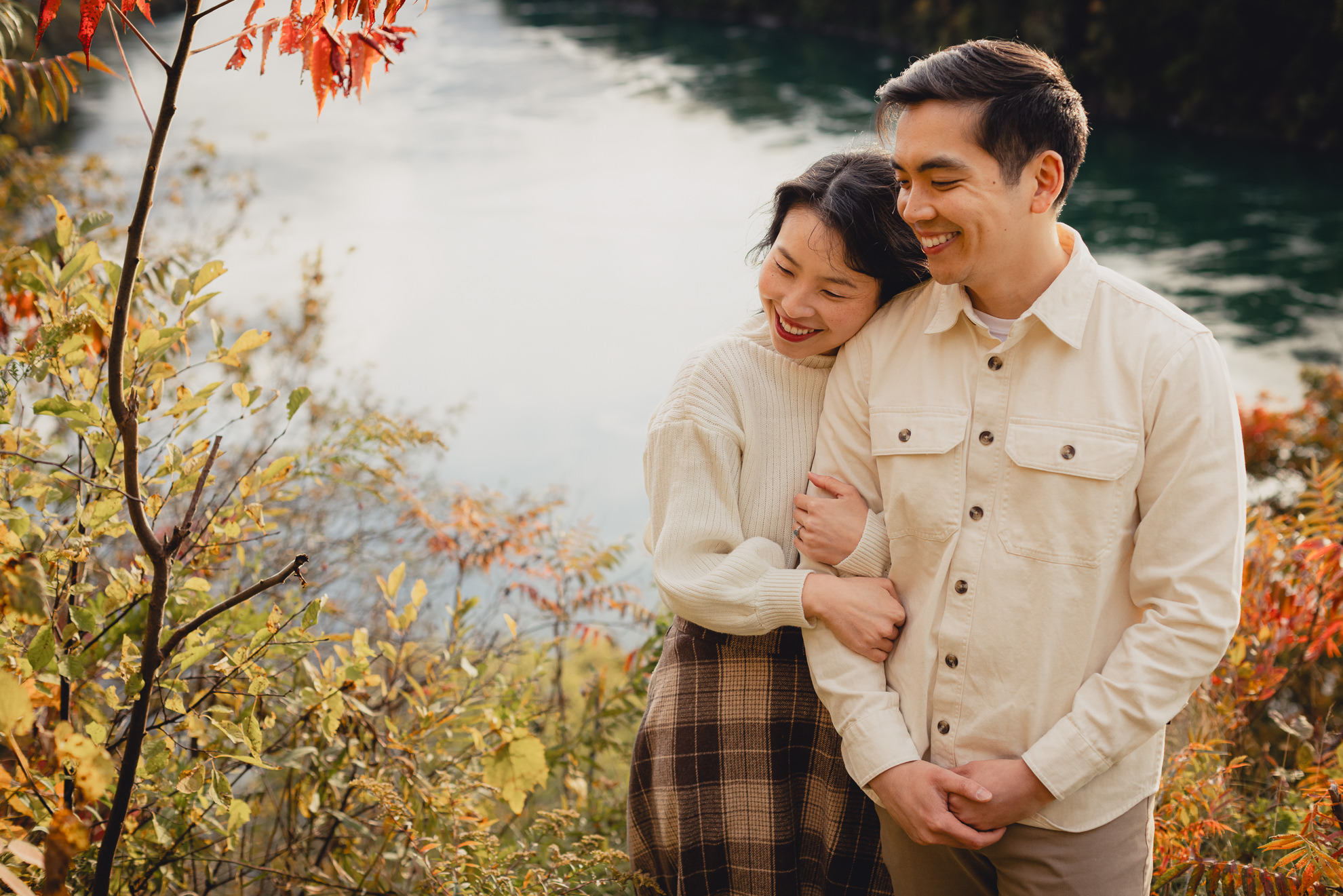 bride hugs grooms arm with fall foliage and river in background during wedding engagement photography session in Niagara Falls, NY