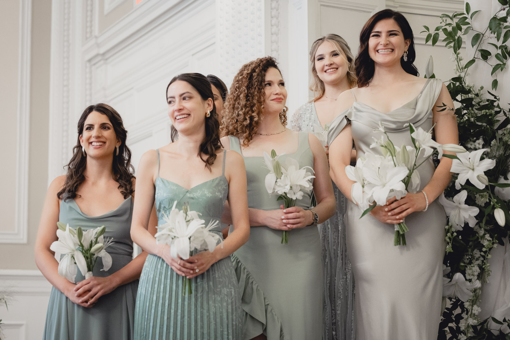 bridesmaids smile during wedding ceremony while holding white lilies