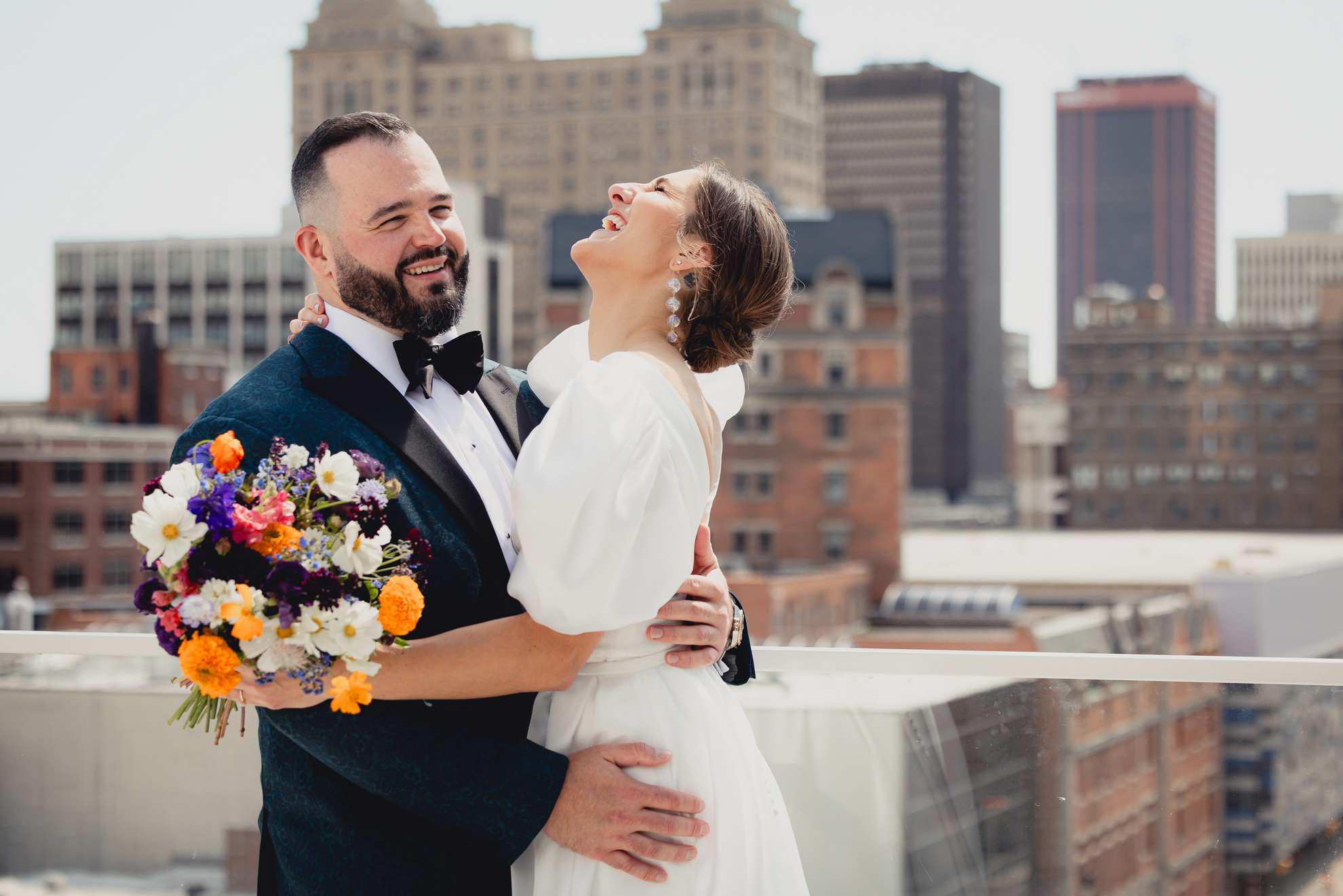 bride and groom embrace and laugh with skyline in background before wedding at Vue on the roof of the Curtiss Hotel in Buffalo, NY
