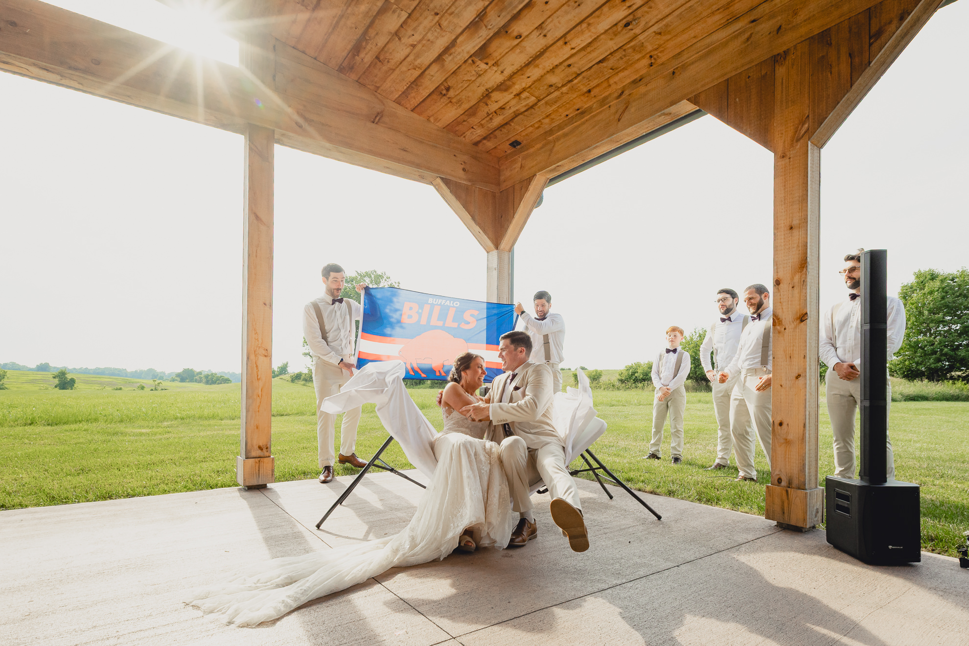 bride and groom jump through folding table in front of Buffalo Bills flag during wedding ceremony at The Gallagher Barn in Medina, NY