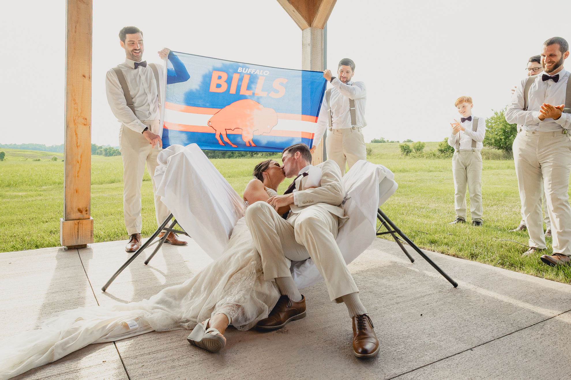 bride and groom kiss after jumping through folding table in front of Buffalo Bills flag during wedding ceremony at The Gallagher Barn in Medina, NY