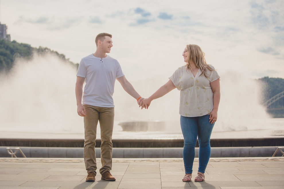 engagement photography of couple holding hands and looking at each other with fountain behind them in Point Park in Pittsburgh