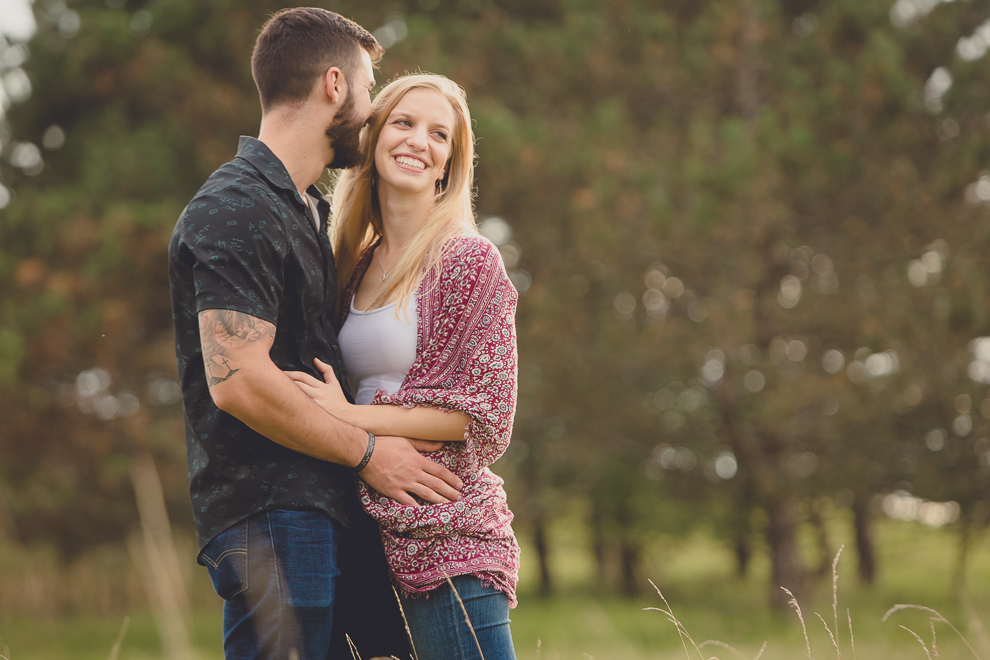 engagement photography of bride laughing as groom kisses her cheek in front of pine trees in field at Tifft Nature Preserve in Buffalo, NY
