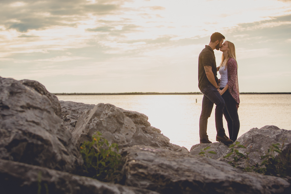 bride and groom kiss on rocks while the sun sets behind them during wedding engagement photography session on Gallagher Beach at Buffalo Harbor