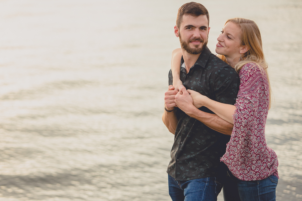 bride wraps arms around grooms shoulders from behind him during wedding engagement photography session on Gallagher Beach at Buffalo Harbor