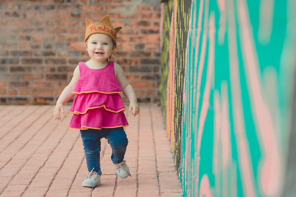little girl wears rebel hat while smiling for photographer during family portrait photography session in front of murals on Chandler St. in Buffalo, NY
