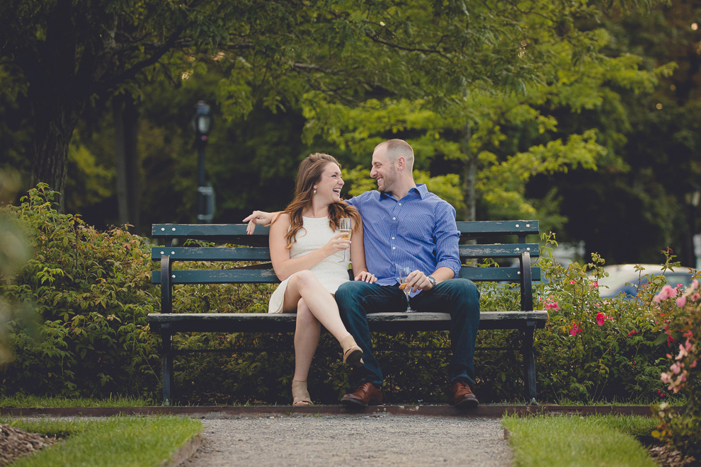 engaged couple laughs on bench in rose garden with champagne glasses for wedding engagement photography at hotel henry & delaware park