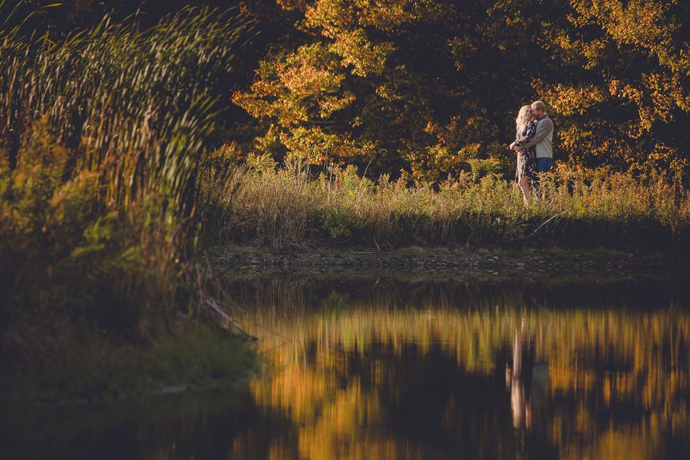 bride and groom hug next to reflective pond during wedding engagement photos among fall foliage at their property in Ellicottville, NY