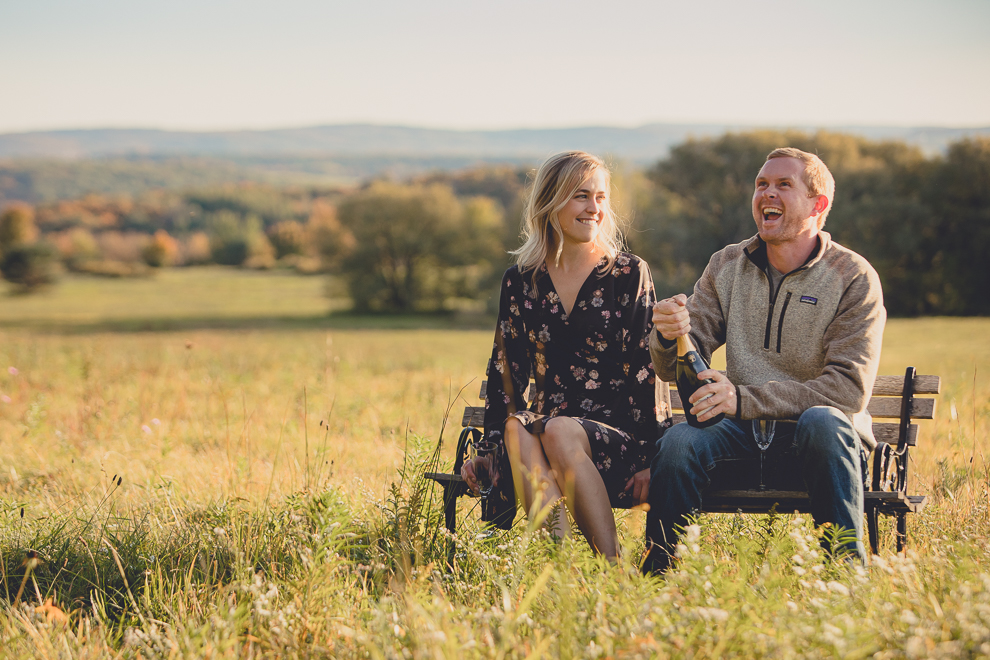 bride and groom enjoy champagne on bench in field during wedding engagement photos among fall foliage at their property in Ellicottville, NY