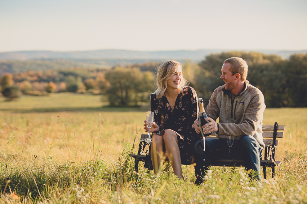 bride and groom enjoy champagne on bench in field during wedding engagement photos among fall foliage at their property in Ellicottville, NY
