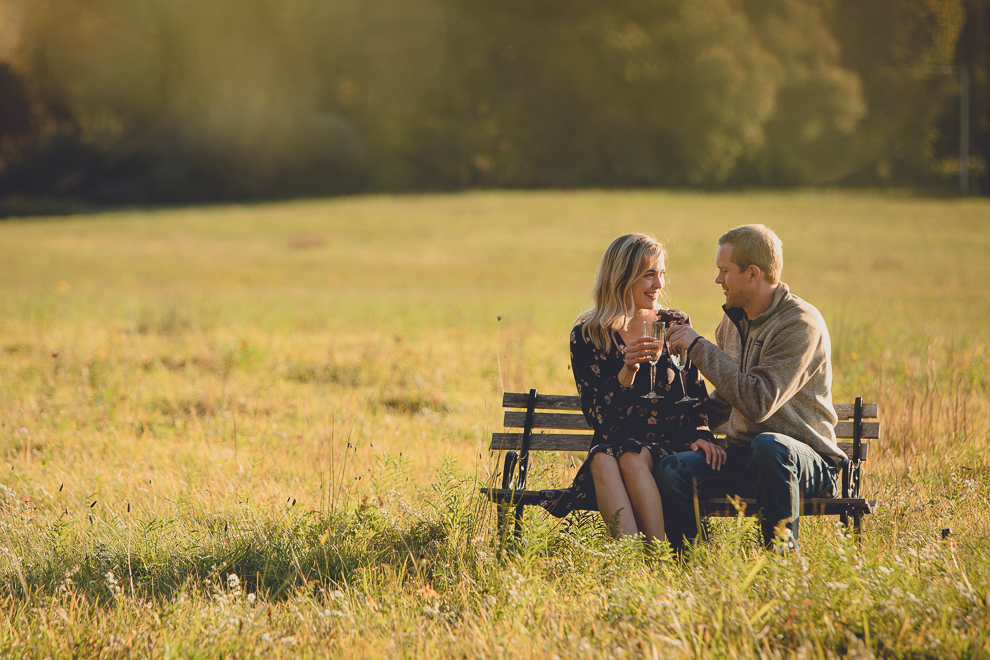 bride and groom enjoy champagne on bench in field during wedding engagement photos among fall foliage at their property in Ellicottville, NY