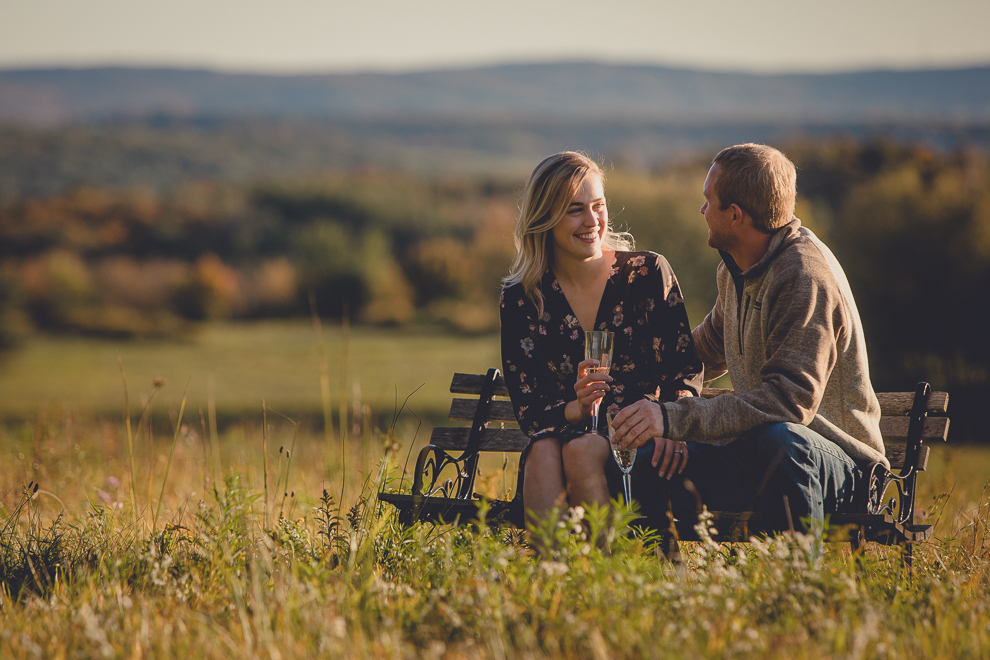 bride and groom enjoy champagne on bench in field during wedding engagement photos among fall foliage at their property in Ellicottville, NY