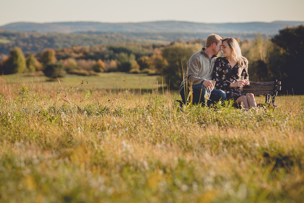 bride and groom enjoy champagne on bench in field during wedding engagement photos among fall foliage at their property in Ellicottville, NY