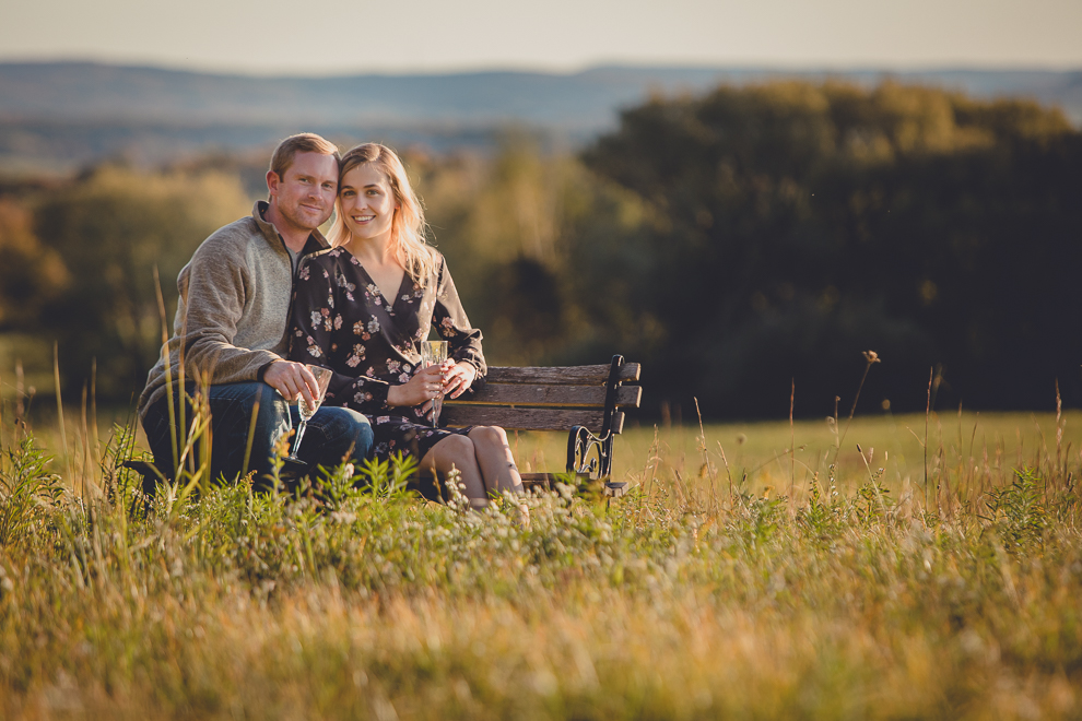 bride and groom enjoy champagne on bench in field during wedding engagement photos among fall foliage at their property in Ellicottville, NY