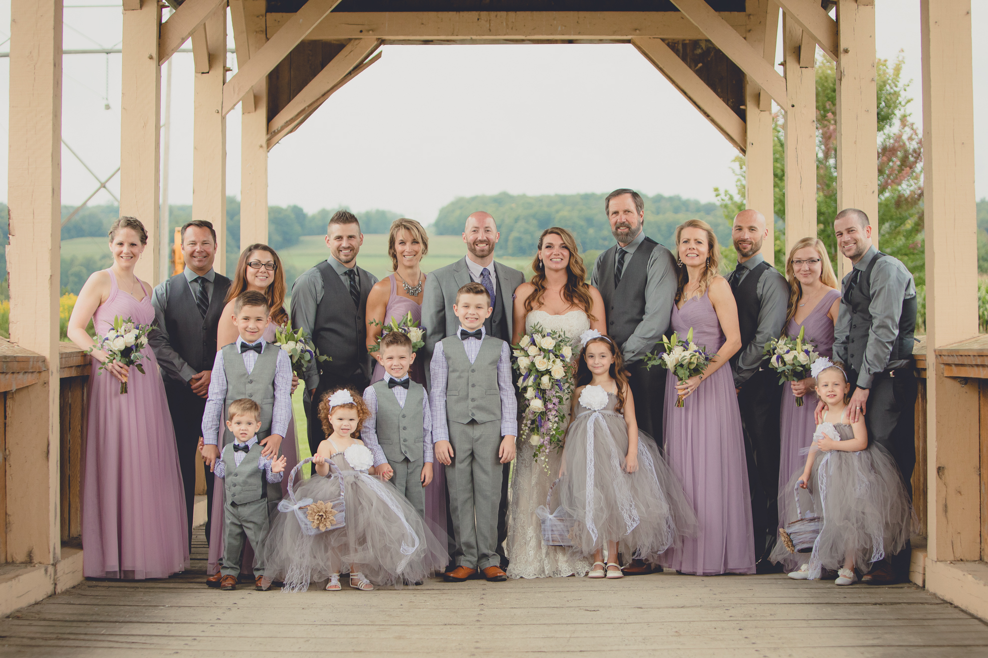 wedding party with flower girls and ring bearers pose for portrait on covered bridge at wedding at hidden valley animal adventure near Buffalo, NY 