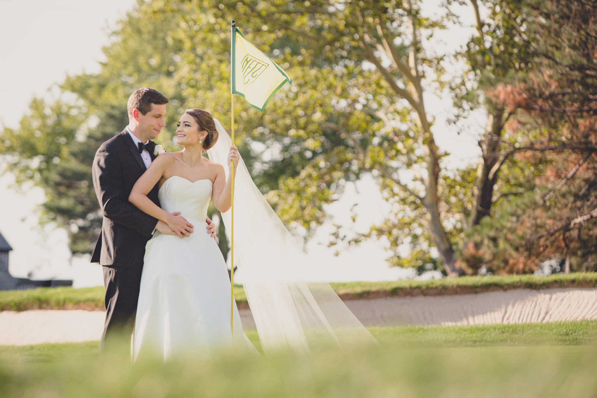 Bride and groom smile at each other next to the flag on the 18th green before wedding reception at Niagara Falls Country Club near Buffalo, NY