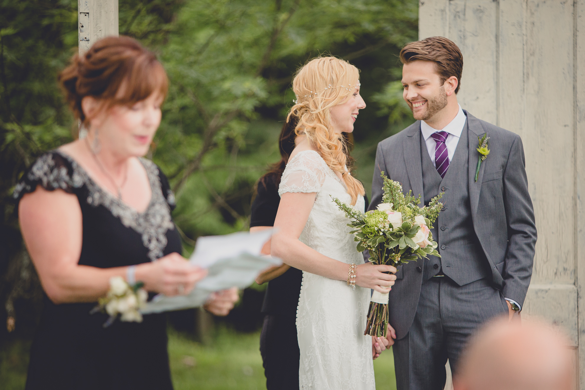 bride and groom smile at each other while mother of bride while she reads during ceremony at small wedding at Knox Farm near Buffalo, NY