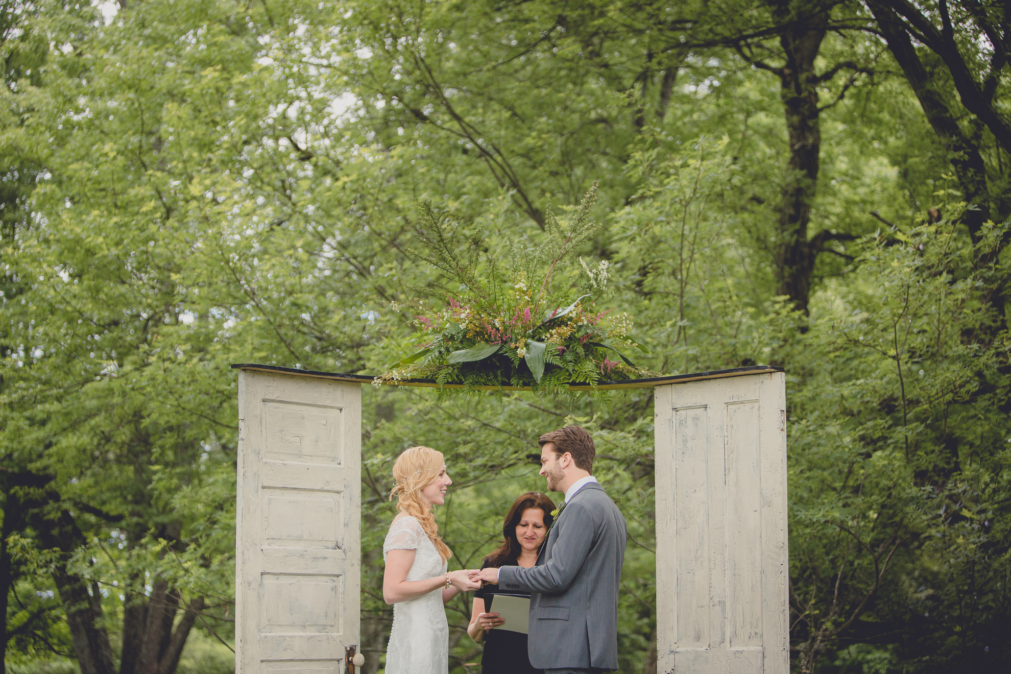 bride places ring on grooms finger between two doors with forest behind them during ceremony at small wedding at Knox Farm near Buffalo, NY