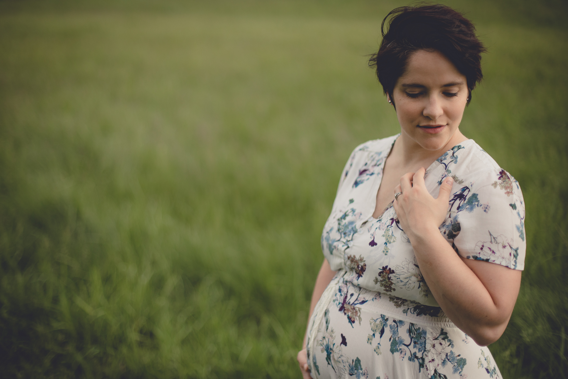 mom looks off to side while wearing floral flowing dress during maternity portrait photography session in field at Tifft Nature Preserve in Buffalo, NY