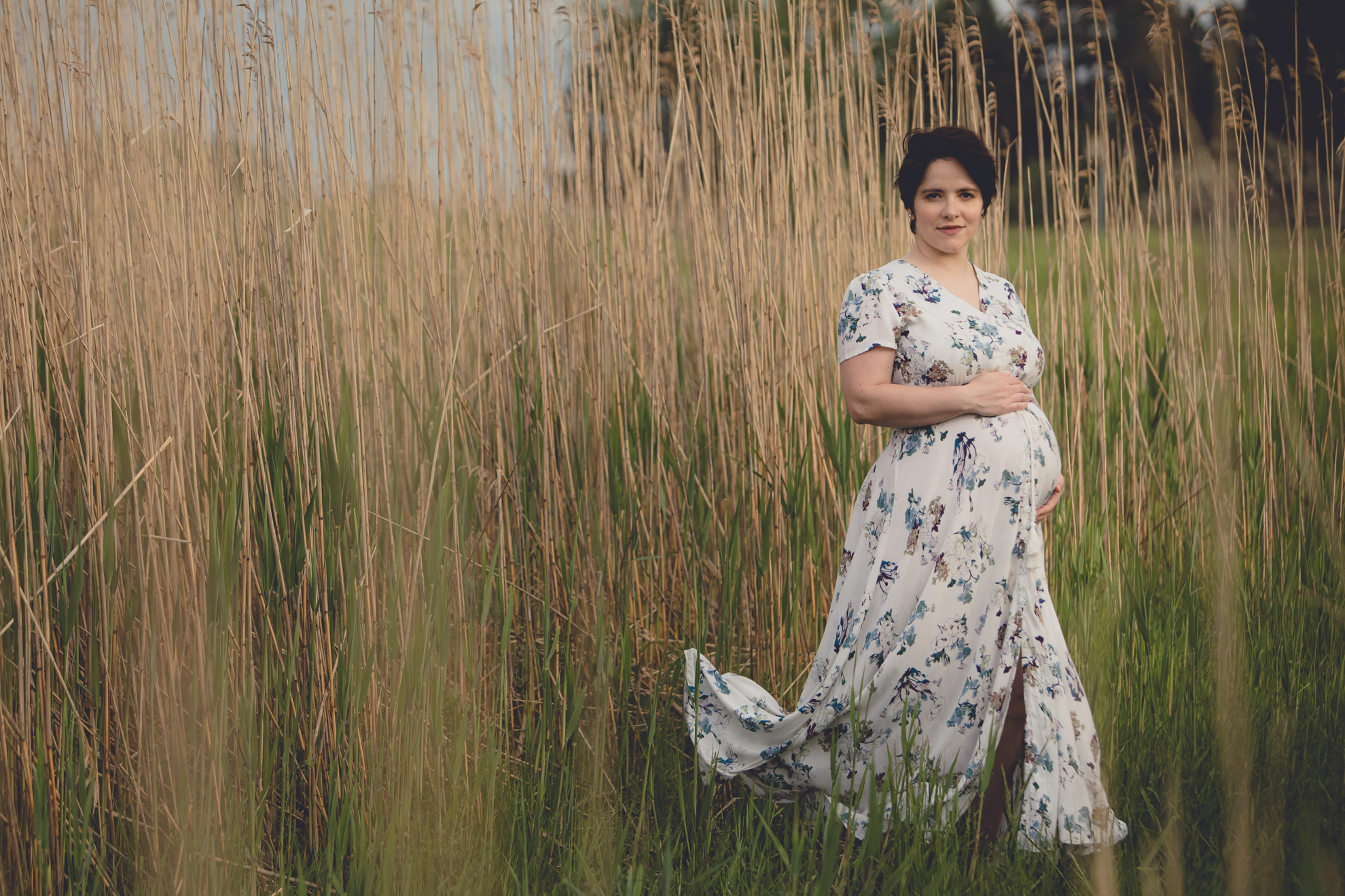mom looks at photographer while holding pregnant stomach during maternity portrait photography session in field at Tifft Nature Preserve in Buffalo, NY