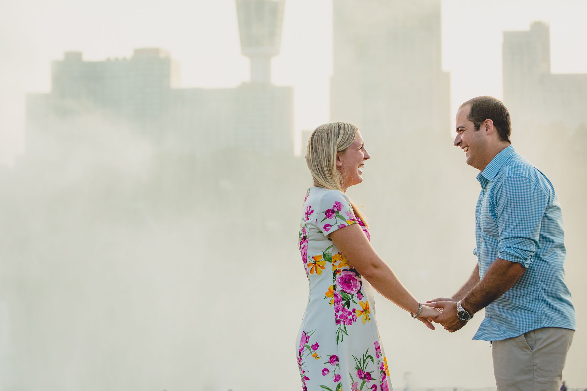 engaged couple holds hands and laughs with Niagara Falls mist and skyline in the background during their wedding engagement photography session 