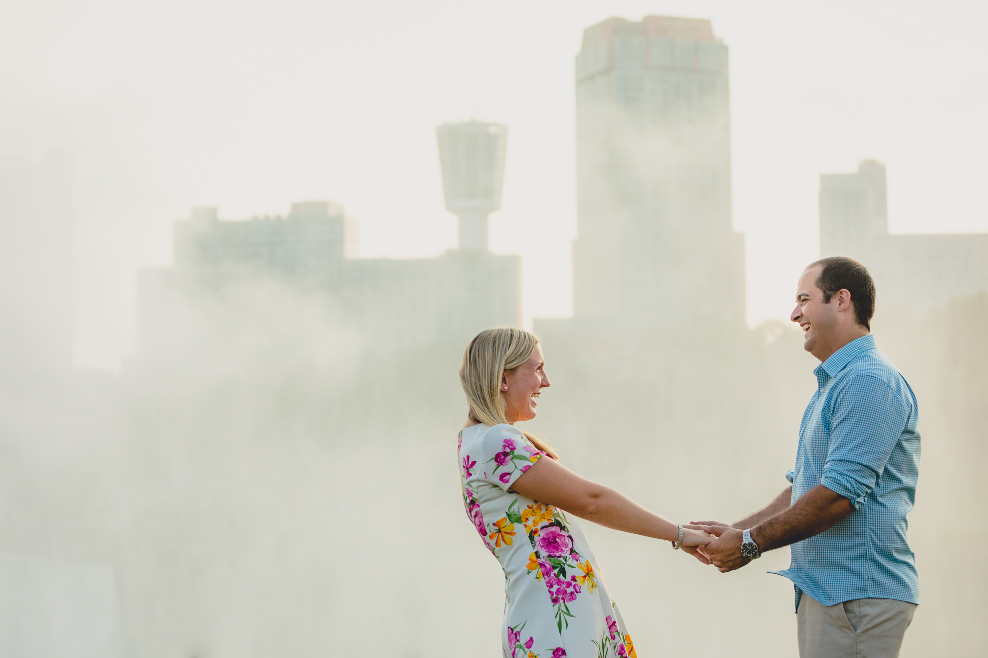 engaged couple holds hands and laughs with Niagara Falls mist and skyline in the background during their wedding engagement photography session 