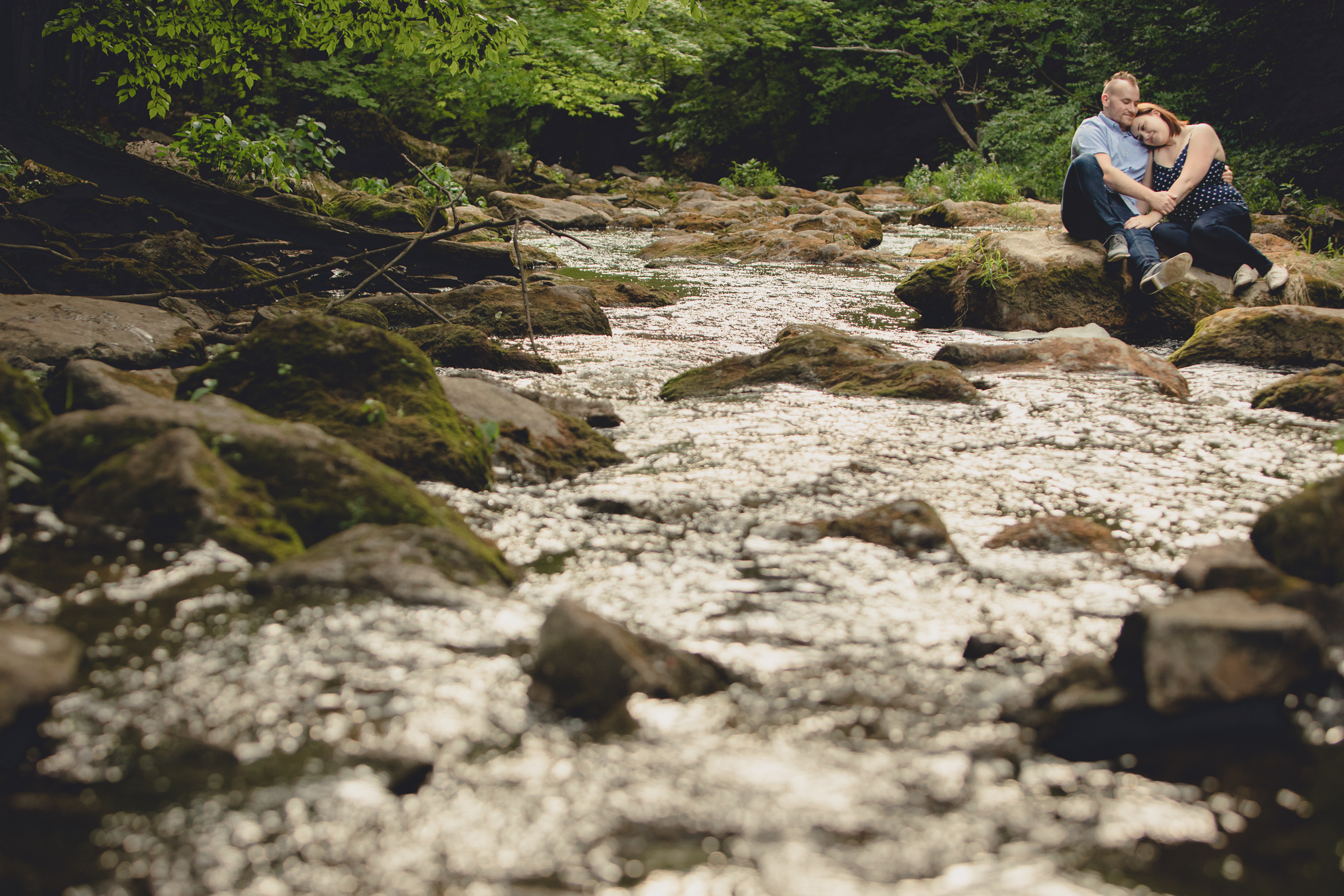 engaged couple cuddles on rock in creek at Akron Falls Park near Buffalo, NY