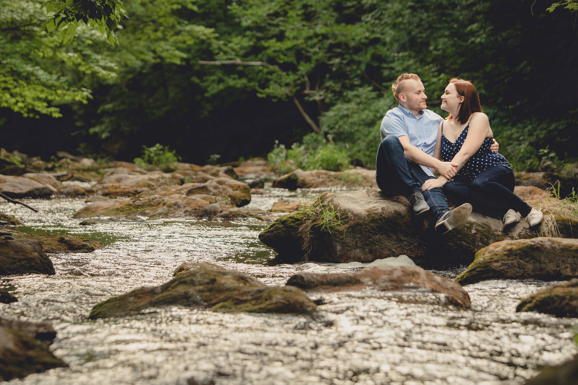 engaged couple looks at each other while sitting on rock in creek during their wedding engagement photography session at Akron Falls Park near Buffalo, NY