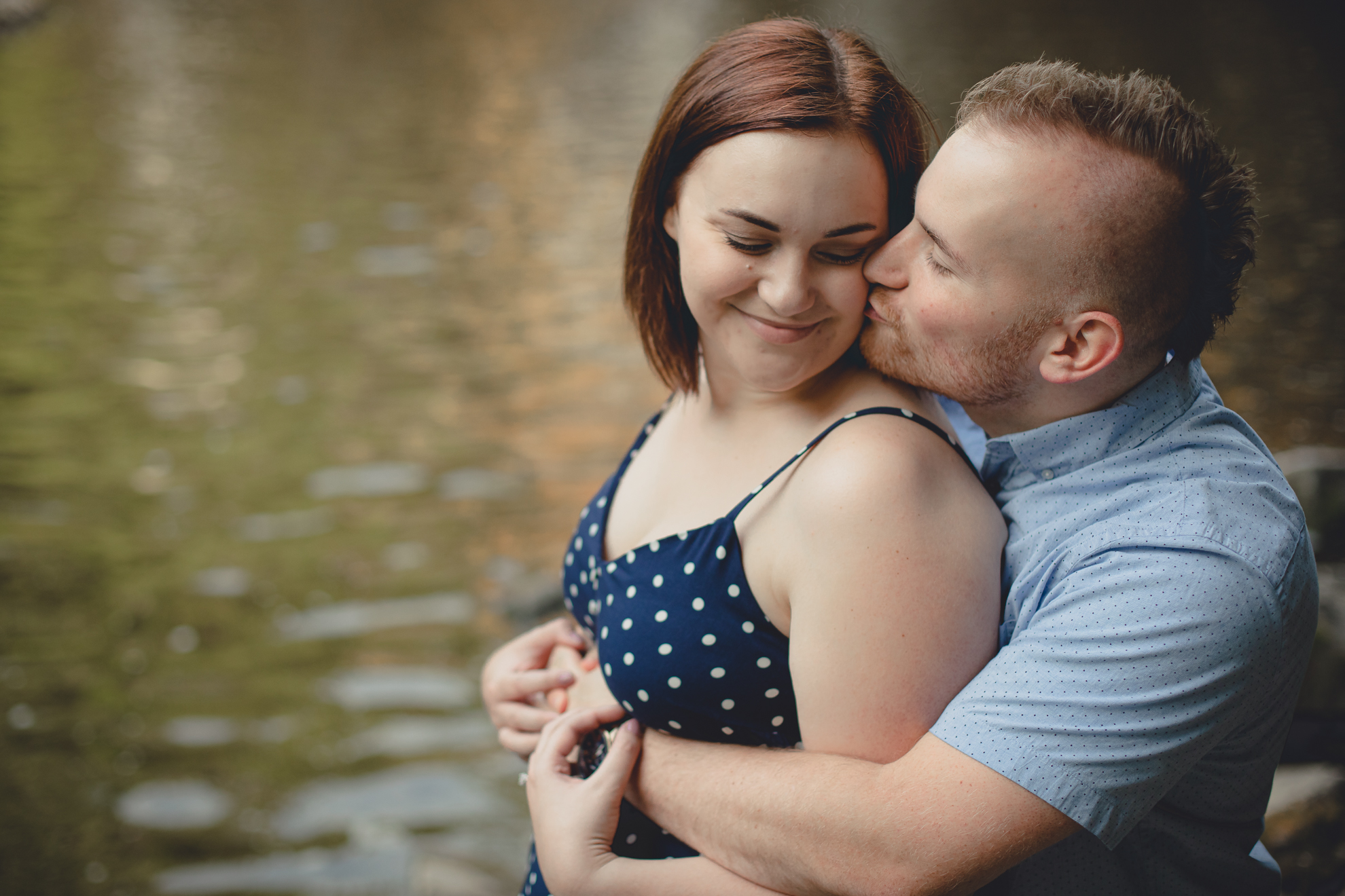 groom kisses bride on cheek in front of waterfall during their wedding engagement photography session at Akron Falls Park near Buffalo, NY