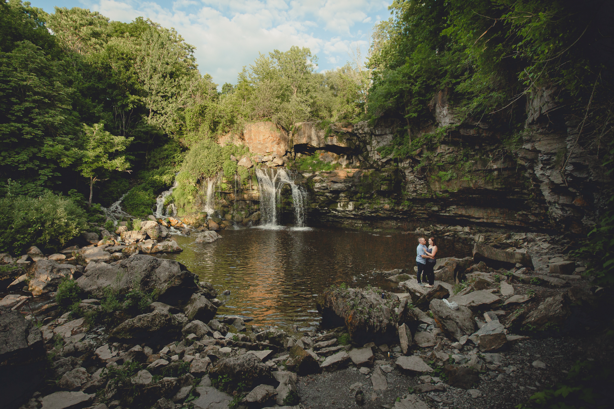 bride and groom stand on rock in front of waterfall during their wedding engagement photography session at Akron Falls Park near Buffalo, NY