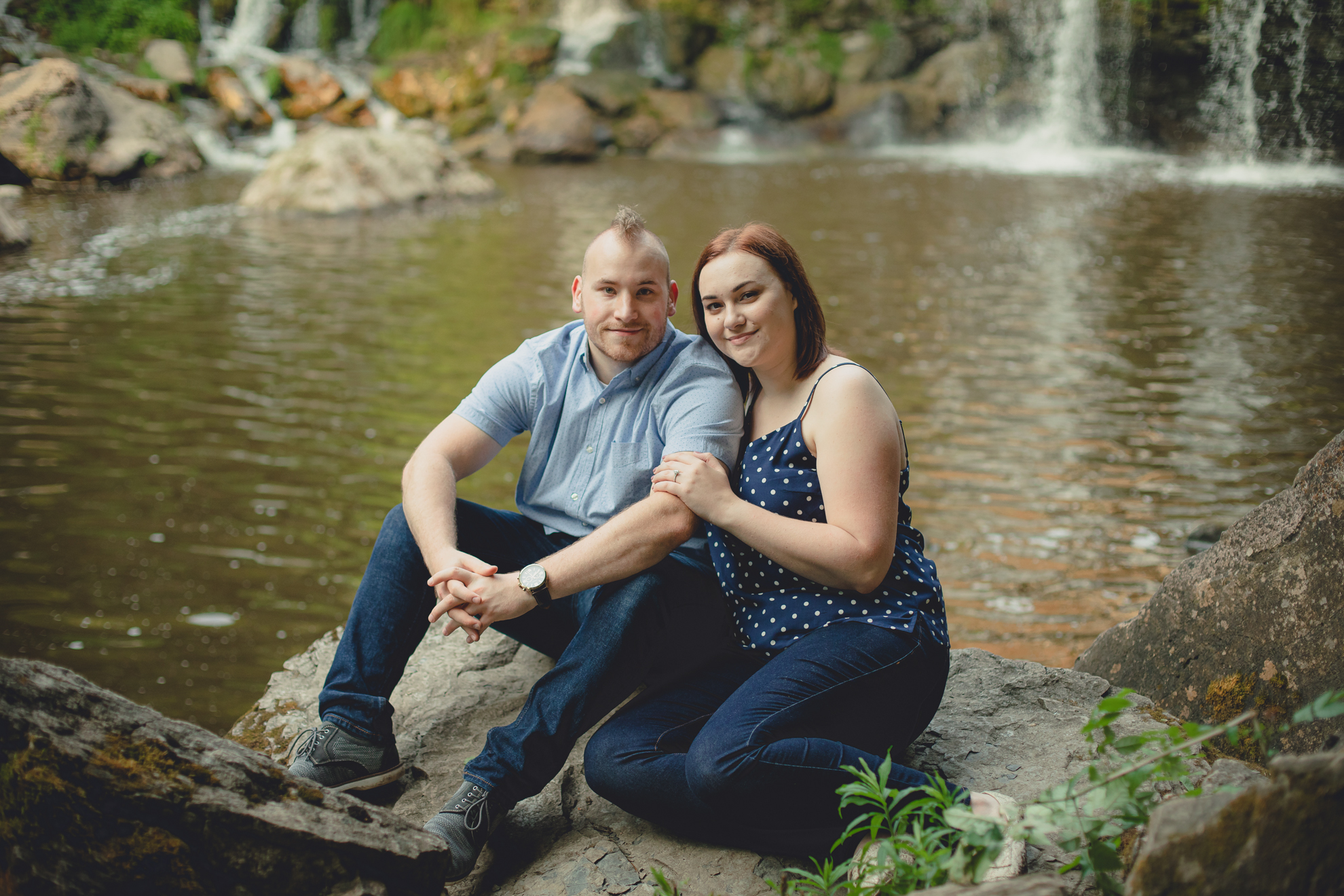 bride and groom sit on rock in front of waterfall smiling for photographer during their wedding engagement photography session at Akron Falls Park near Buffalo, NY