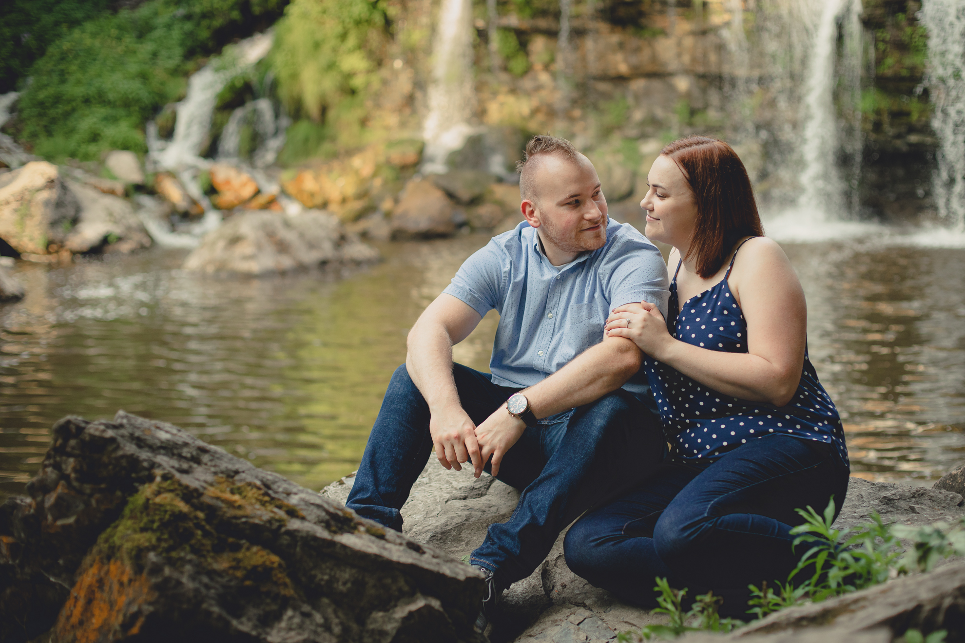 bride and groom sit on rock in front of waterfall during their wedding engagement photography session at Akron Falls Park near Buffalo, NY
