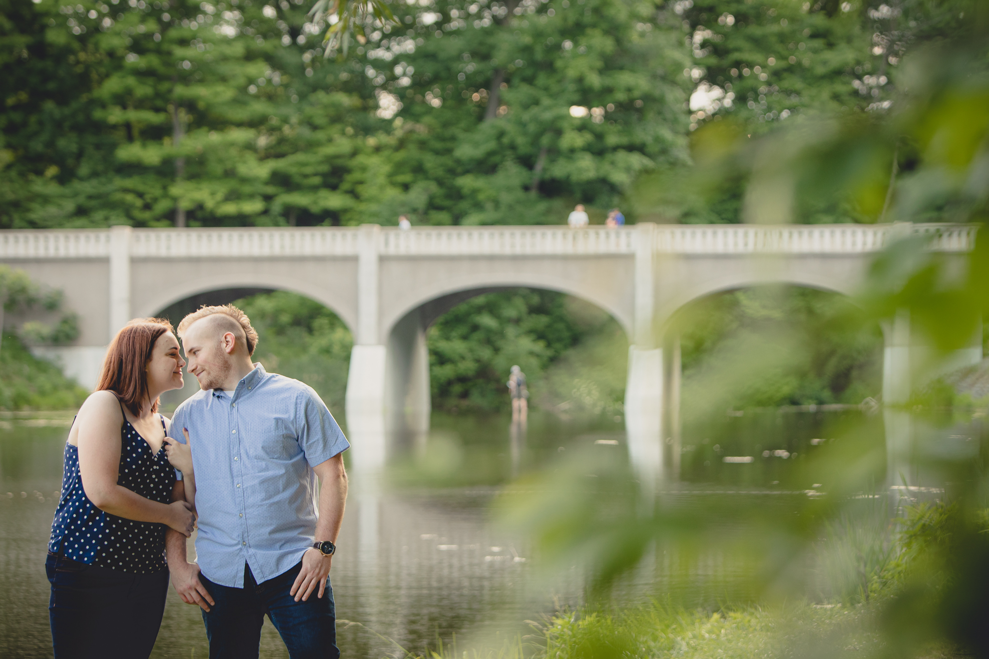 engaged couple poses for photographer in front of bridge and lake at Akron Falls Park near Buffalo, NY