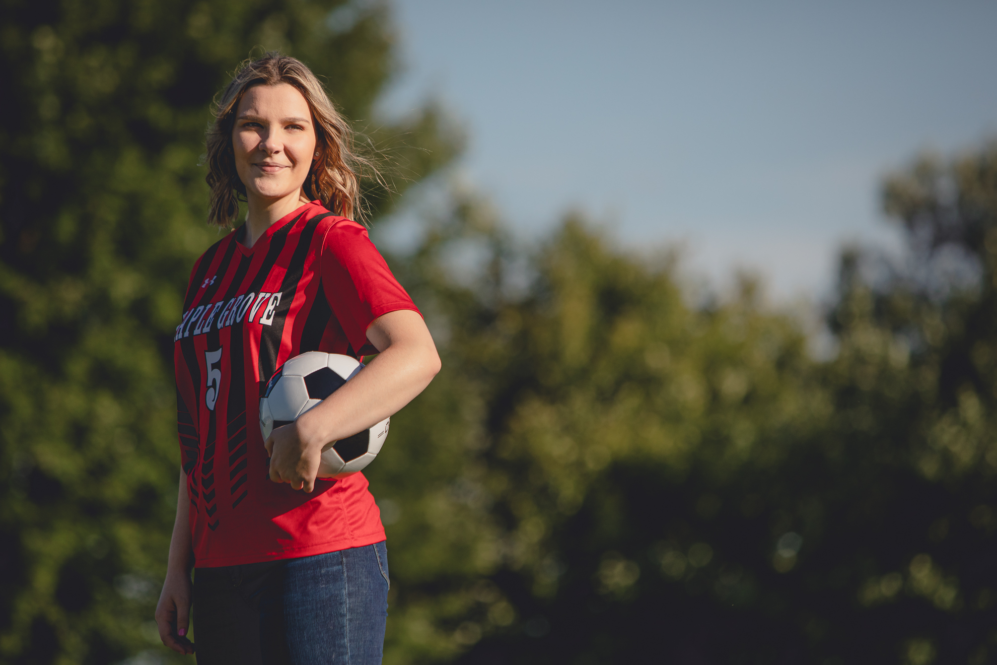 maple grove high school student smiles poses for photographer while wearing jersey in soccer field during senior portrait photography session at Chautauqua Institution