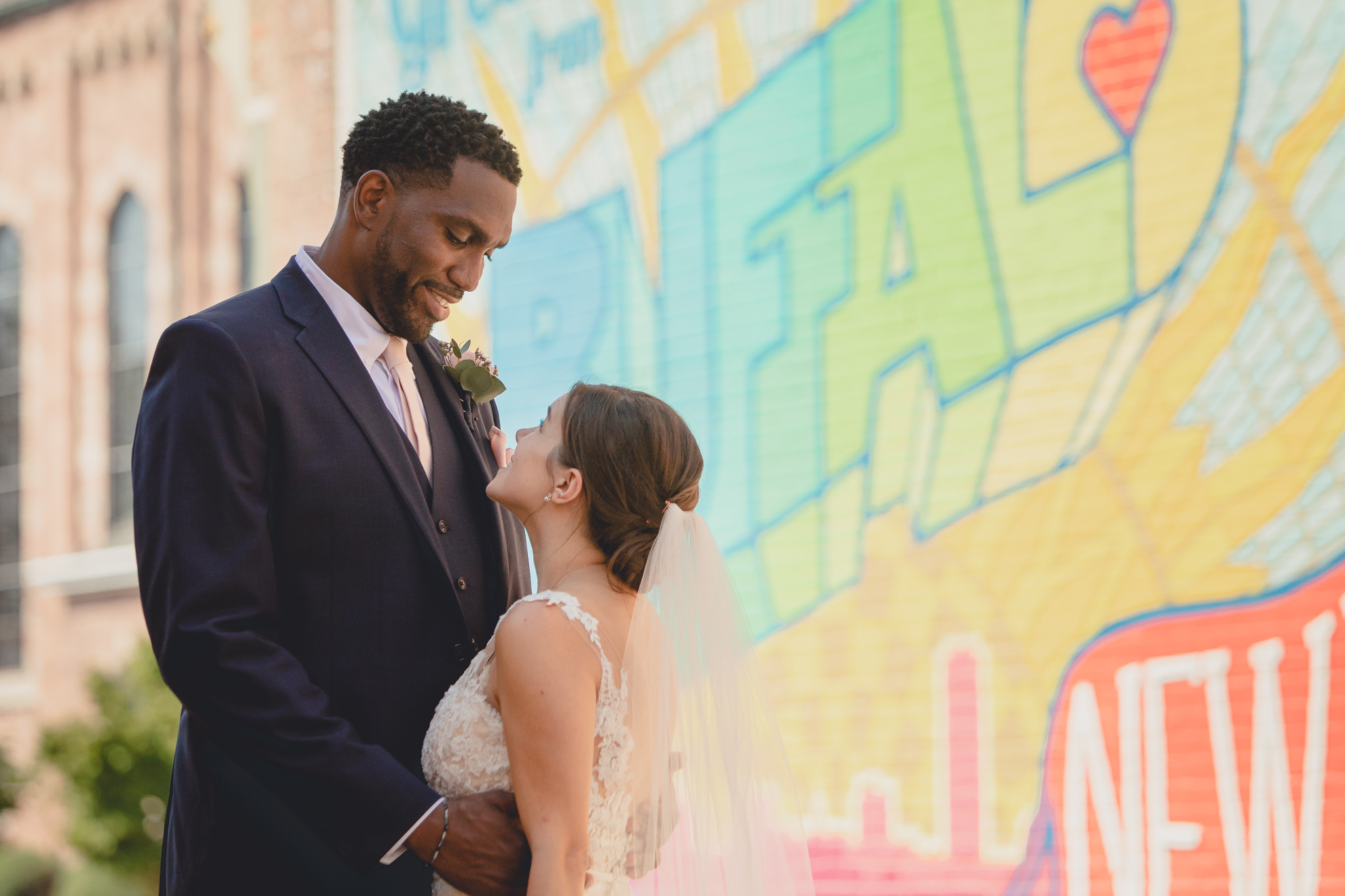 bride and groom smile at each other in theater district of Buffalo, NY at the "greetings from Buffalo" mural during WNY micro wedding celebration