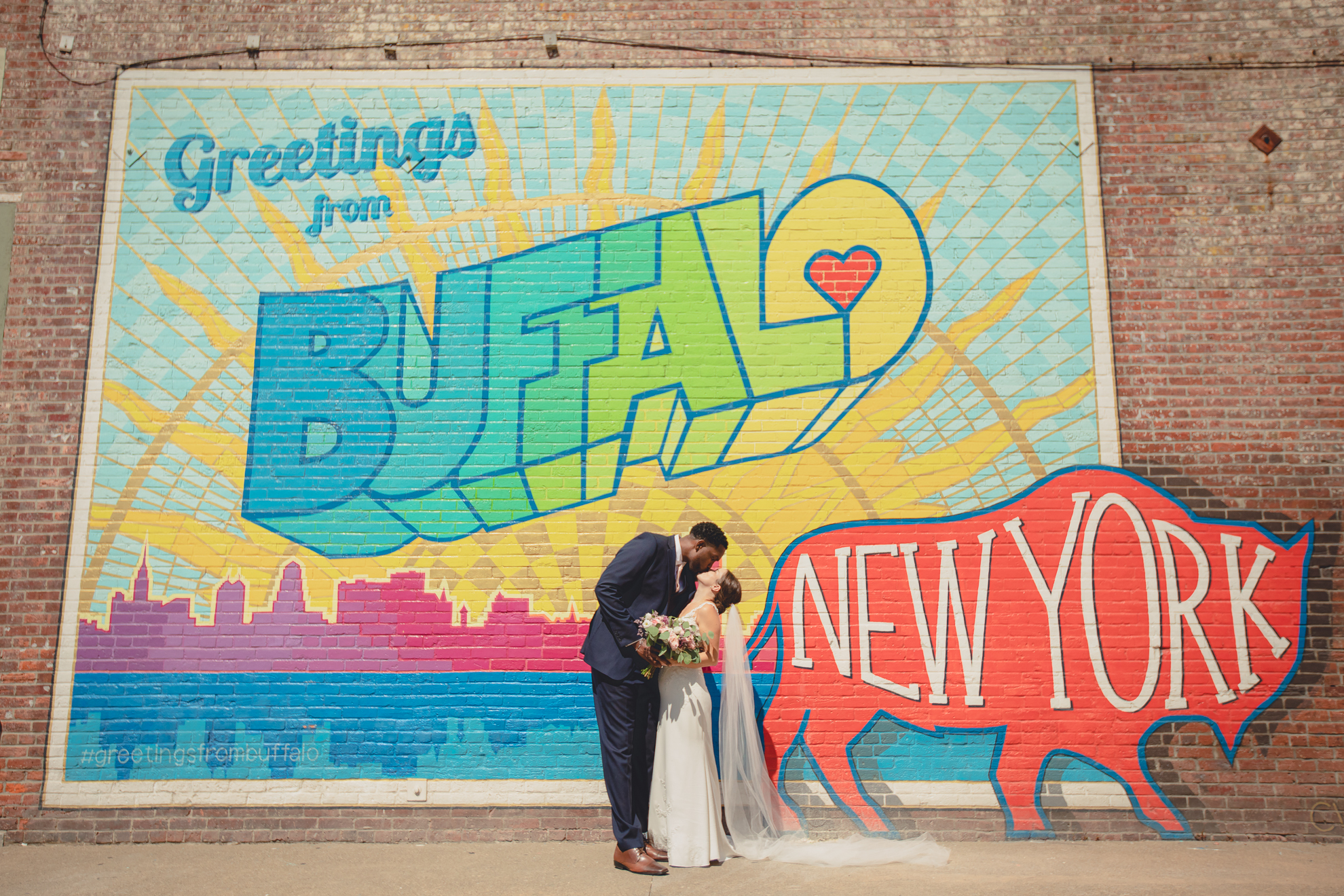 bride and groom kiss in theater district of Buffalo, NY at the "greetings from Buffalo" mural during WNY micro wedding celebration