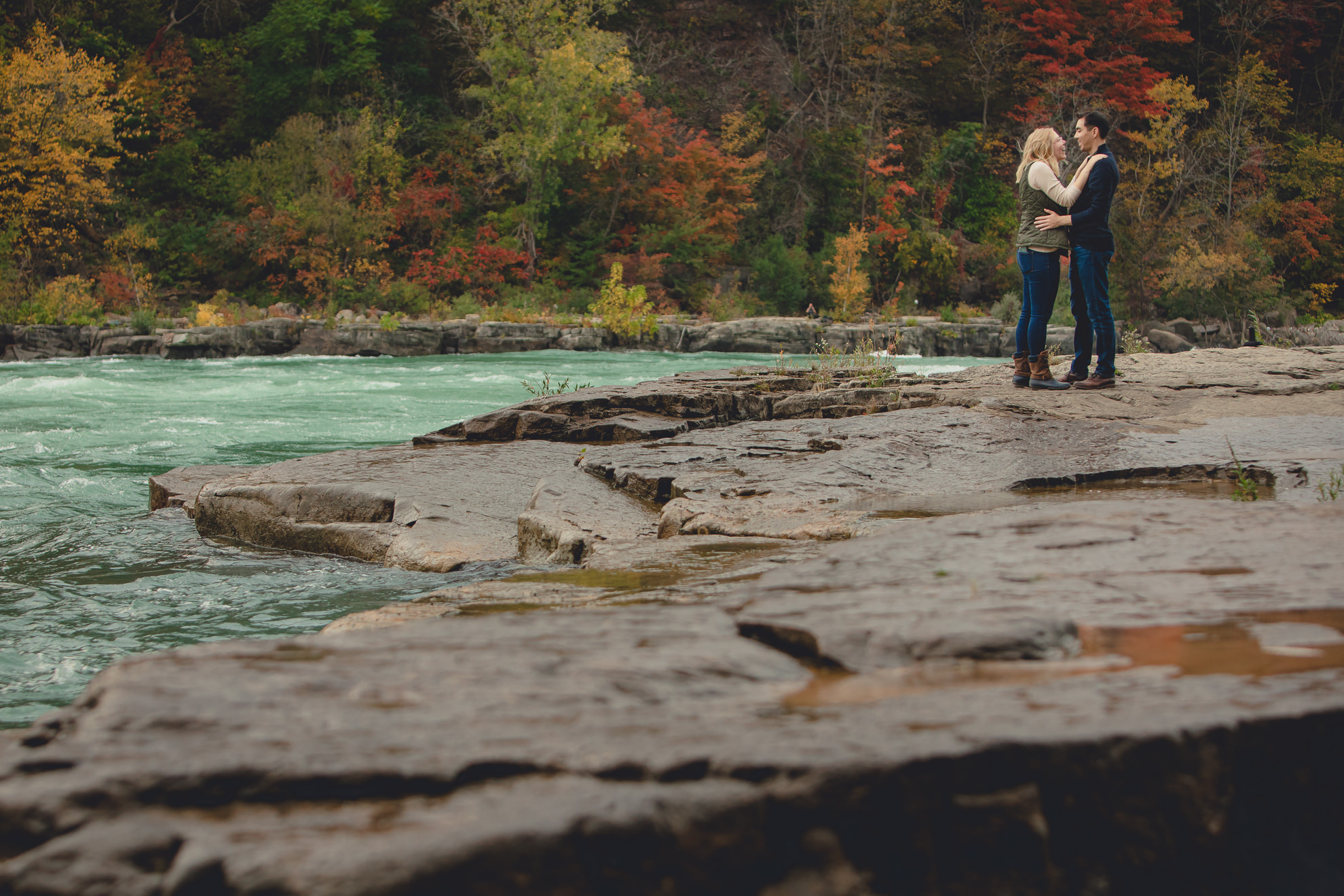 couple hugs after wedding engagement proposal photography with rapids and fall foliage in background at Whirlpool State Park in Niagara Falls, NY