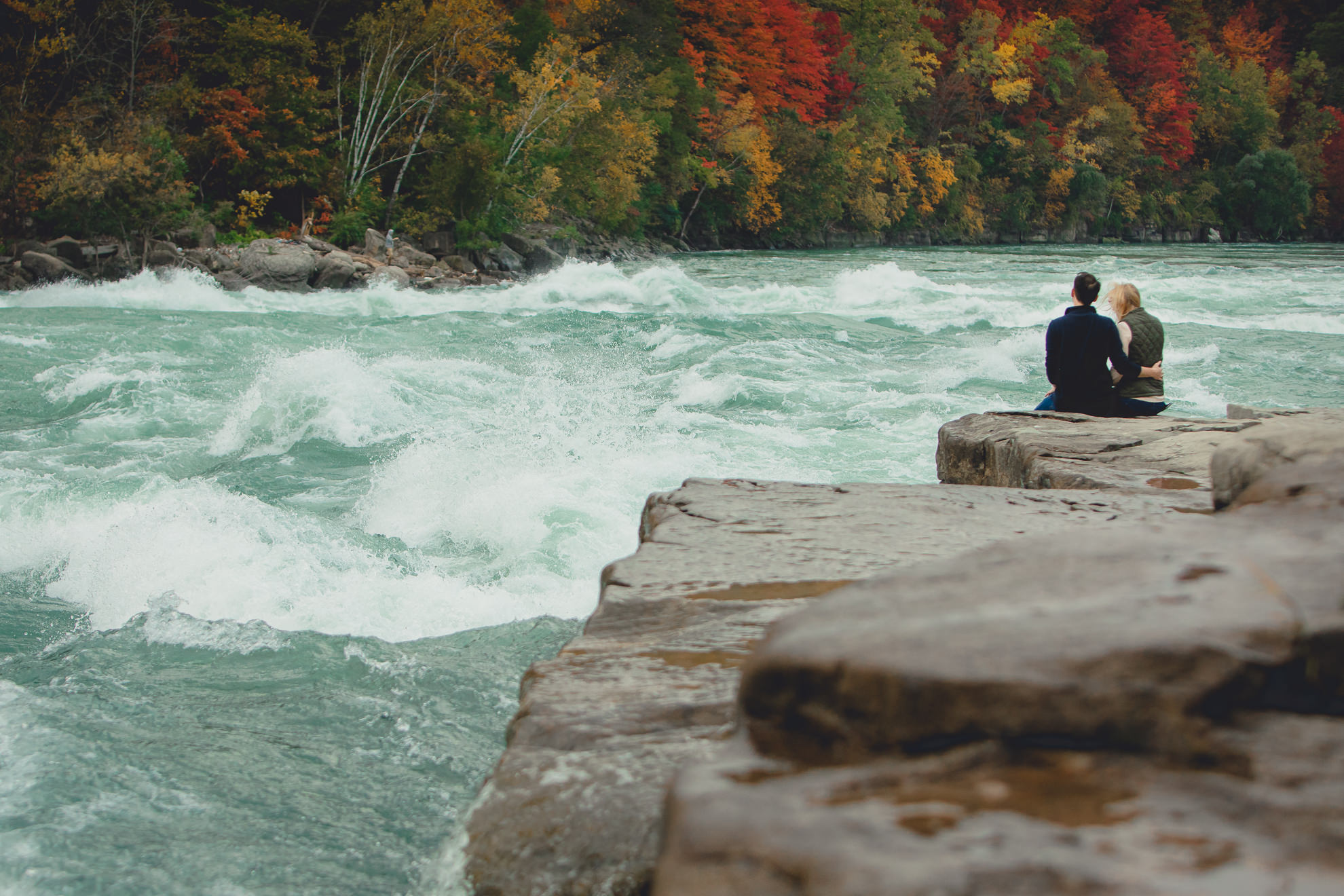 couple sits and admires rapids on rock after wedding engagement proposal photography with rapids and fall foliage in background at Whirlpool State Park in Niagara Falls, NY