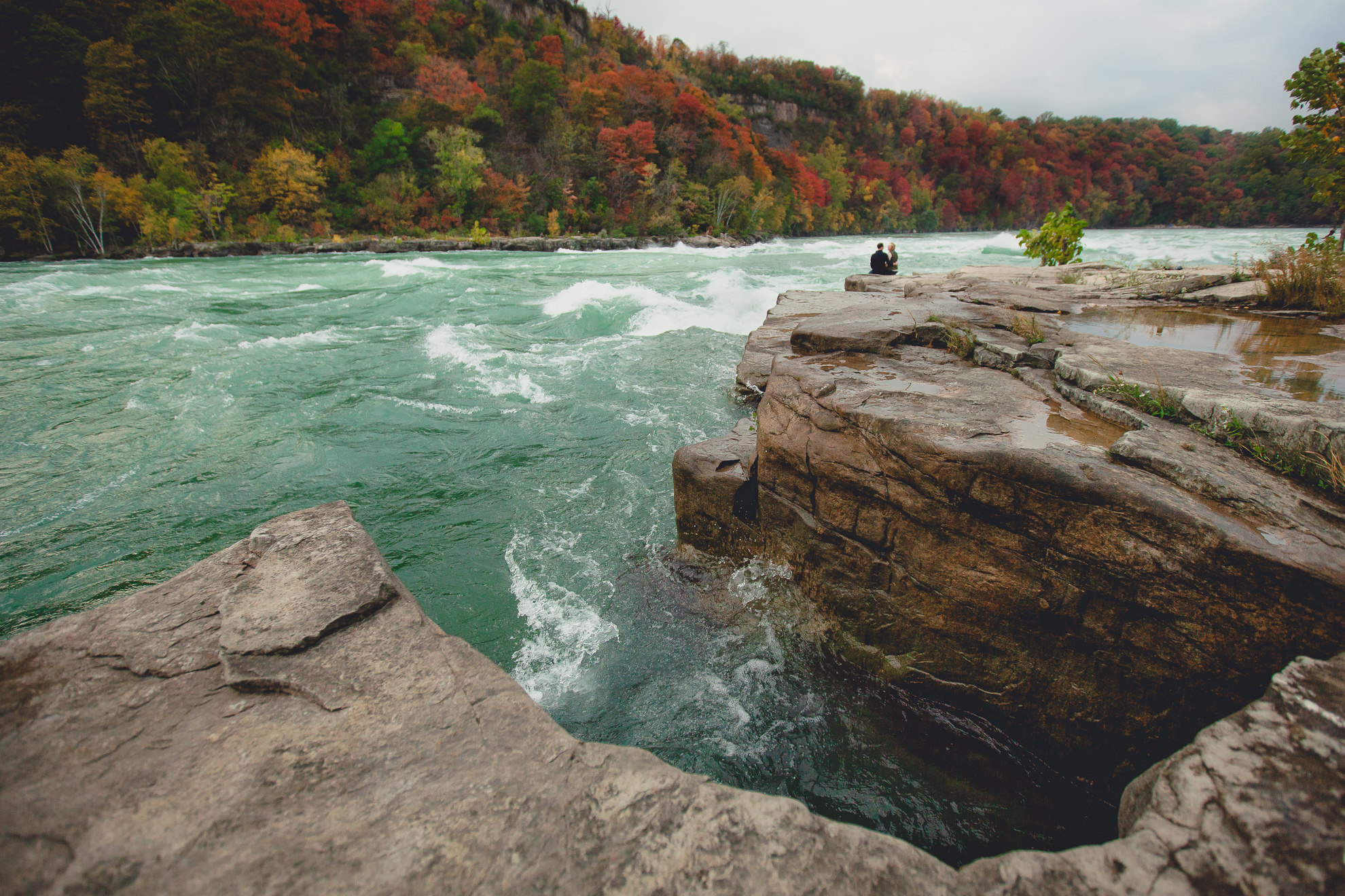 couple sits and admires rapids on rock after wedding engagement proposal photography with rapids and fall foliage in background at Whirlpool State Park in Niagara Falls, NY