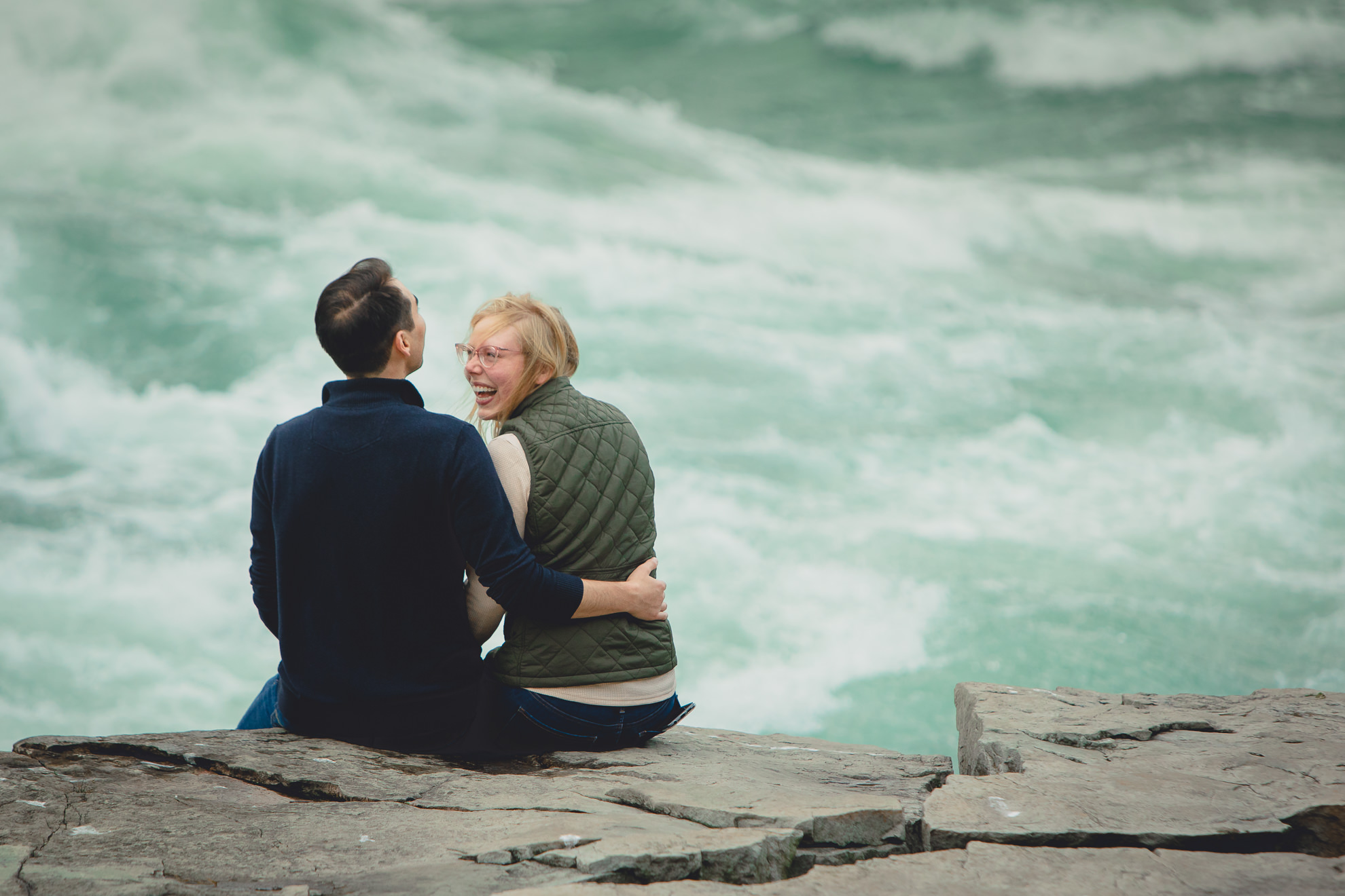 couple sits and admires rapids on rock after wedding engagement proposal photography with rapids and fall foliage in background at Whirlpool State Park in Niagara Falls, NY